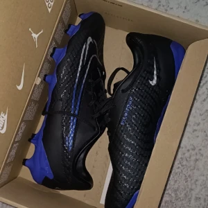 Svarta och blåa fotbollsskor från Nike. Köpt för 1200 sälj för 1100  - Säljer ett par svarta fotbollsskor från Nike med blå detaljer och vit Nike-logga. Skorna har snörning och är designade för att ge bra grepp på planen. Perfekta för dig som vill ta ditt spel till nästa nivå. 