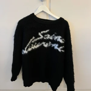 Svart stickad tröja från Saint Laurent - Säljer en svart stickad tröja från Saint Laurent med vit, abstrakt text på framsidan. Tröjan har lång ärm och en lite fluffig, mjuk struktur. Perfekt för dig som gillar exklusiva och trendiga plagg.storlek M helt nytt skick. 