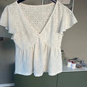 Vit blus - Söt vit blus med v-ringning och korta volangärmar. Blusen har broderade hål i blommönster och en lätt utsvängd passform. Perfekt för varma dagar och ger en romantisk känsla.