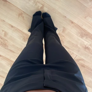 Svarta kostymbyxor med normal passform - Säljer ett par svarta kostymbyxor med klassisk rak passform och hög midja. Byxorna har diskreta fickor bak och bälteshällor i midjan. Perfekta för en stilren och enkel look. Även slits vid fötterna🥰