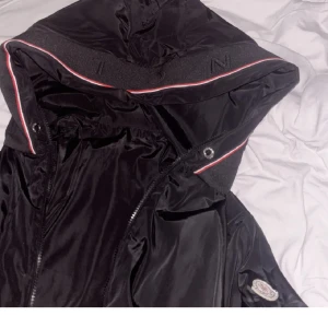 Svart windbreaker från Moncler med huva - Säljer en svart windbreaker från Moncler med huva och dragkedja. Jackan har Moncler-logga på ärmen och detaljer i rött och vitt längs huvan. Perfekt för dig som vill ha en stilren och sportig look.