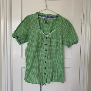 Grön rutig skjortblus med spetsdetalj - Säljer en grön och vit rutig blus från Orbis Trachten med korta puffärmar, knappar framtill, vit spets vid ringningen och snörning i ryggen. Köpt vintage. S/M
