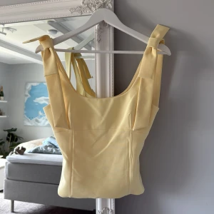 Gul topp med breda axelband från Zara - Säljer en ljusgul topp från Zara med breda axelband som knyts på axlarna. Toppen har en figurnära passform och är tillverkad i ett slätt, mjukt material. Perfekt för vår och sommar! Använd endast 1 gång och säljer på grund av att den tyvärr var för stor på mig.