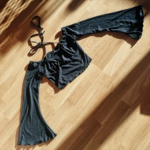 Svart croppad topp med knytband - Trendig svart croppad topp med långa ärmar och smala knytband som knyts som en halter-topp tillsammans med säkerhets nålar som även korsas på framsidan. Toppen har en tight passform och är tillverkad i ett ribbat material som ger en snygg struktur.