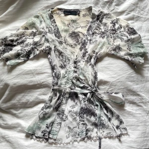 Vit och svart blus med knytband - Säljer en tunn och luftig blus från Kappahl i vitt med svart mönster. Blusen har knytband i midjan, v-ringning, knappar framtill och korta ärmar. Perfekt för dig som gillar romantisk och feminin stil.