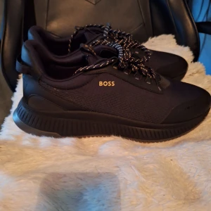 Svarta sneakers från BOSS - Säljer ett par svarta sneakers från BOSS med snygga svarta och vita snören. Skorna har en sportig design med rund tå och platt sula. Ovandelen är i textil och syntet, och BOSS-loggan syns i guld på sidan. Helt oavnända