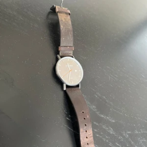 Klassisk herrklocka från Sidegren behövs byte på batteri  - Stilren klocka från Sidegren med rund silverfärgad urtavla, datumvisning och brunt skinnarmband. Urtavlan har minimalistisk design med tydliga markeringar och orange sekundvisare. Perfekt accessoar för dig som gillar enkel och tidlös stil.
