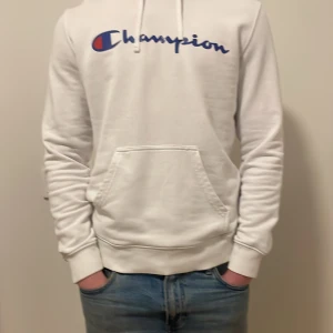Vit hoodie från Champion - Säljer en klassisk vit hoodie från Champion med blå och röd logga på bröstet. Tröjan har huva med dragsko och en stor magficka. Perfekt för en avslappnad stil.