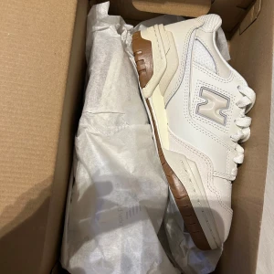 Beige New Balance 550 sneakers - Säljer ett par stilrena New Balance 550 sneakers i beige med vita och ljusbruna detaljer. Skorna har klassisk siluett, perforerade paneler och gummisula i brunt. Perfekta för dig som gillar en clean och sportig look. Helt nya ur förpackning missade tyvärr returdagen dem blev lite väll små för mig. Hör av dig för fler bilder eller så eventuella frågor 🤗