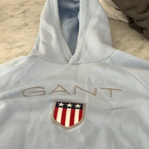 Ljusblå hoodie från GANT - Säljer en ljusblå hoodie från GANT med broderad logga och sköld på bröstet. Tröjan har huva och en stor magficka framtill. Perfekt för dig som gillar klassisk och sportig stil.