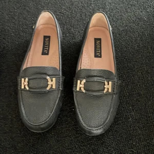 Svarta loafers från Novita med gulddetaljer - Stilrena svarta loafers från Novita med guldfärgad metallspänne framtill. Skorna har rund tå och är tillverkade i skinn med en klassisk design. Perfekta för en snygg och tidlös look. Strl 39 använda en gång pga storlek. 