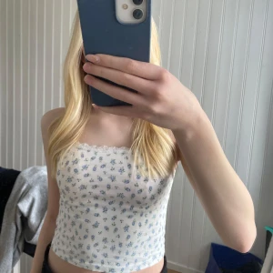 Vit blommig linnetopp med spets - Söt vit linnetopp med små blå blommor och grönbladigt mönster. Toppen har smala axelband och en fin spetskant upptill. Perfekt för varma dagar och enkel att matcha med jeans eller kjol.
