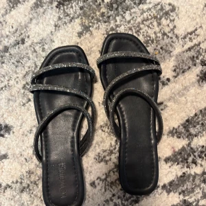 Svarta sandaler med glittriga remmar - Säljer ett par svarta sandaler med fyrkantig tå och tre smala remmar dekorerade med glittriga stenar. Perfekta för att lyfta en outfit med lite extra bling. Snygg och trendig design.