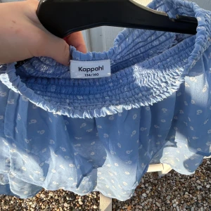 Blå blommig blus från Kappahl - Söt blå blus från Kappahl med små vita blommor och volangdetaljer. Blusen har en smockad resår upptill och är tillverkad i ett lätt och luftigt material. Perfekt för varma dagar!