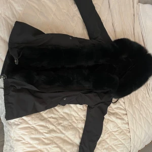 Svart parkajacka med päls - Säljer en svart parkajacka med stor, fluffig päls längs huva, krage och framtill. Jackan har dragsko i midjan och nederkant, samt långa ärmar. Perfekt för kyliga dagar och ger en lyxig känsla till outfiten.