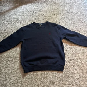 Mörkblå sweatshirt från Polo Ralph Lauren - Klassisk mörkblå sweatshirt från Polo Ralph Lauren med rund halsringning och diskret röd broderad logga på bröstet. Tröjan har långa ärmar och ribbade muddar vid ärmslut och nederkant. Perfekt för en stilren och avslappnad look.