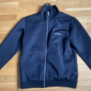 Svart jacka med dragkedja från Les Deux - Säljer en svart jacka från Les Deux med hög krage och hel dragkedja framtill. Jackan har två fickor på sidorna och diskret logga på bröstet. Enkel och stilren design som passar till många tillfällen.