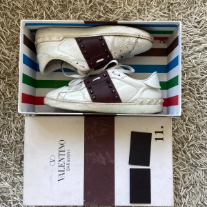 Valentino Garavani sneakers med nitar - Säljer ett par vita sneakers från Valentino Garavani med bred mörkröd rem över mitten dekorerad med nitar. Skorna har rund tå, platt sula och snörning. Klassisk och stilren design med exklusiv känsla.