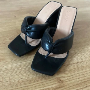 Svarta klackade sandaler med knut - Säljer ett par svarta sandaler med breda remmar och snygg knutdetalj framtill. Skorna har fyrkantig tå och hög klack, vilket ger en trendig look. Helt oanvända. Köpta från Asos 