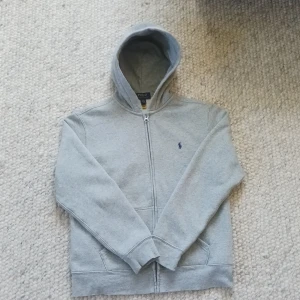 Grå hoodie från Polo Ralph Lauren - Säljer en klassisk grå hoodie från Polo Ralph Lauren med dragkedja och huva. Tröjan har en broderad marinblå logga på bröstet och två fickor framtill. Perfekt för en avslappnad stil.