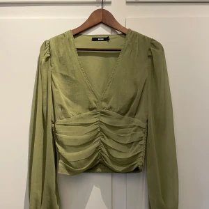 Olivgrön blus med v-ringning och rynkade detaljer - Säljer en grön blus från Bik Bok med djup v-ringning och rynkade detaljer framtill. Blusen har långa, lätt transparenta ärmar och smock baktill i midjan. Perfekt för dig som gillar trendiga och feminina plagg.