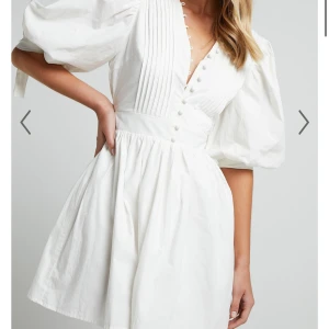 Vit kortklänning med puffärmar och knappar - Superfin vit klänning med puffiga trekvartsärmar och knytband vid ärmslut. Djup v-ringning med dekorativa knappar framtill och veckade detaljer upptill. Perfekt för studenten, aldrig använd med lapp kvar 