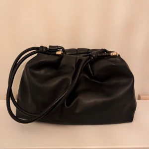 Svart axelväska - Säljer en svart axelväska i skinnimitation med snygga gulddetaljer. Väskan har ett rymligt huvudfack och en innerficka med dragkedja. Perfekt för dig som vill ha en stilren och trendig accessoar.