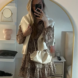 Beige leopardmönstrad klänning med volang - Säljer en beige klänning med leopardmönster och volangdetaljer. Klänningen har långa ärmar och en lös passform, perfekt för dig som gillar trendiga och bekväma plagg. Matchas snyggt med en stickad tröja över axlarna och en vit axelväska.