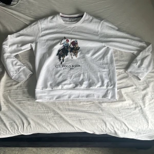 Vit sweatshirt från U.S. Polo Assn. - Säljer en vit sweatshirt från U.S. Polo Assn. med tryck av två polospelare på framsidan. Tröjan har rund halsringning och långa ärmar. Klassisk och stilren design med broderad logga och mjukt material. Jättefin. Knappt använd.