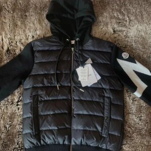 Svart cardigan jacka med huva från Moncler - Säljer en svart cardigan jacka från Moncler med vadderad framsida, stickade ärmar och huva. Jackan har dragkedja, två fickor fram och en vit detalj på ena ärmen. Perfekt för dig som vill ha en snygg och sportig look.