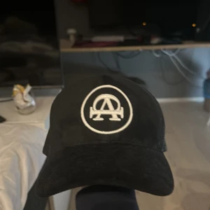 Svart keps från All Cap one - Svart keps med vit broderad logga framtill och texten 'All Cap One' broderad på sidan. Kepsen har mesh på sidorna och justerbar snapback baktill. Perfekt för en stilren och avslappnad look.