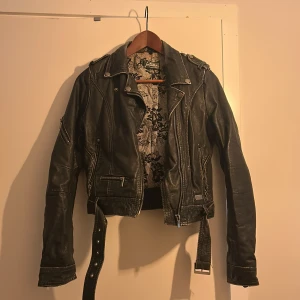 Svart skinnjacka från Jofama by Kenza - Säljer en svart skinnjacka från Jofama by Kenza med coola detaljer som dragkedjor, axelklaffar och bälte i midjan. Jackan har ett snyggt mönstrat innerfoder och klassisk bikerstil. Perfekt för dig som vill ha en edgy look. Denna köptes för ca 2000kr och har lite skavanker. 