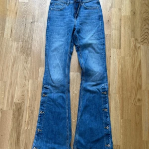 Blå bootcut jeans med knappar längs benen - Säljer ett par blå bootcut jeans med dekorativa knappar längs utsidan av benen. Jeansen har klassisk femficksdesign och en snygg tvätt. Perfekta för dig som vill ha en unik detalj på dina jeans. (Dock lite slitna baktill i nedkant pga lång modell och använda av 160 samt 168 cm lång)