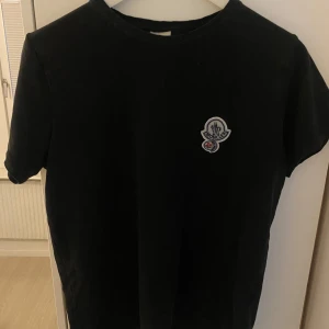 Svart t-shirt från Moncler - Säljer en svart t-shirt från Moncler med rund halsringning och korta ärmar. På bröstet finns en liten broderad Moncler-logga i vitt och rött. Perfekt för en stilren och enkel look.