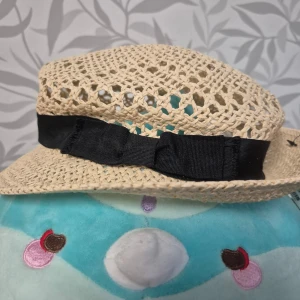 Beige stråhatt med svart rosett H&M S - Snygg beige stråhatt från H&M med bred svart rosett runt kullen. Hatten är flätad och har ett luftigt, öppet mönster som passar perfekt för soliga dagar. Klassisk form och lätt material gör den både stilren och bekväm. Helt ny, oanvänd. 