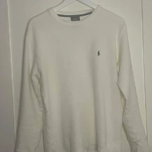 Vit långärmad tröja från Ralph Lauren - Klassisk vit långärmad tröja från Ralph Lauren med den ikoniska broderade loggan på bröstet. Tröjan har rund halsringning och är tillverkad i mjuk bomull, perfekt för en stilren och enkel look.