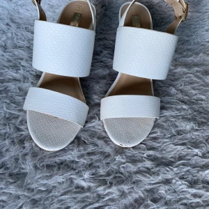 Vita sandaler med klack och strukturmönster - Säljer ett par vita sandaler med breda remmar och strukturmönster. Skorna har en öppen tå och en stabil, brun klack. Perfekta för varmare dagar och ger en stilren look till outfiten.