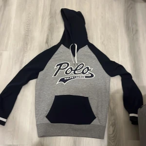 Grå och marinblå hoodie från Polo Ralph Lauren - Säljer en snygg hoodie från Polo Ralph Lauren i grått och marinblått med stor logga framtill. Tröjan har huva med dragsko och en stor magficka. Perfekt för en avslappnad stil.
