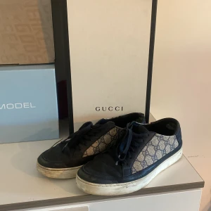Gucci skor - Säljer ett par snygga Gucci sneakers med klassiskt blått och beige GG-monogram på sidorna. Skorna har svarta skinnpartier, snörning och vit platt sula. Perfekta för dig som gillar lyxiga och har gått sönder lite på framsidan av höger skorn du ser de på andra bilden.