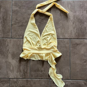 Gul söt topp med volang - Säljer en ljusgul söt topp med knyt i nacken och volang nertill. Toppen är ärmlös och har en croppad passform, perfekt för varma dagar. Storlek 34, normal i storlek. 