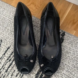 Svarta pumps med rosett och öppen tå - Säljer ett par svarta pumps med låg klack och öppen tå. Skorna har en dekorativ rosett framtill med små metalländar och är tillverkade i en kombination av glansigt och matt material. Perfekta för dig som vill ha en klassisk men ändå unik sko. Kan rengöras innan jag skickar de