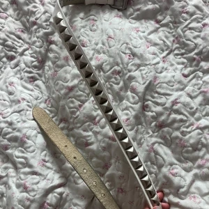 Vitt nitarbälte med silverfärgade pyraminnitar - Säljer ett vitt bälte med silverfärgade pyramidformade nitar längs hela bandet. Bältet har klassisk spänne och är gjort i ett material som liknar skinn. Perfekt accessoar för att ge outfiten en edgy touch.