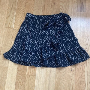 Svart blommig omlottkjol  - Supersöt svart omlottkjol från Newyorker med små vita blommor och volangkant. Kjolen har knytband i midjan och är perfekt för sommaren.