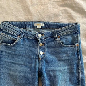 Blå bootcut jeans med knappar - Säljer ett par blå bootcut jeans med synliga knappar framtill. Jeansen har låg midja och klassisk femficksdesign. Justerbara i midjan. Passar mig som e 165🥰🥰