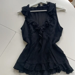 Svart volangblus från H&M - Säljer en svart blus från H&M i storlek 38. Blusen har v-ringning med volangdetaljer längs halsringningen och framtill. Materialet är lätt och har en subtil struktur, perfekt för en trendig look.
