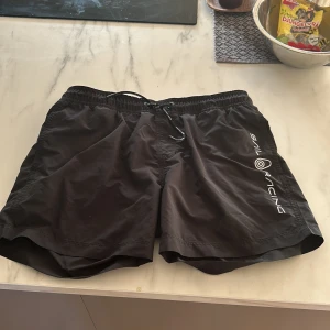 Svarta badshorts från Sail Racing - Svarta badshorts från Sail Racing med vit logga på ena benet. Shortsen har snörning i midjan och är tillverkade i 100% polyester. Perfekta för sommardagar vid vattnet. Knappast använda, nya kostar ungefär 500kr  priset går att diskutera