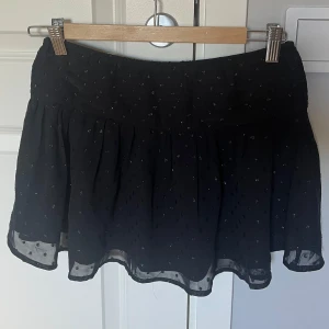 Svart prickig kortkjol från Gina Tricot - Supersöt svart kortkjol från Gina Tricot med glittriga prickar och lätt volang i nederkanten. Storlek 158/164 och är nästan helt oanvänd.