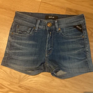 Blå jeansshorts från Replay - Säljer ett par klassiska blå jeansshorts från Replay med fem fickor, knapp och dragkedja framtill. Shortsen har låg midja och är i en normal passform. Perfekta för sommaren och enkla att matcha med olika toppar.