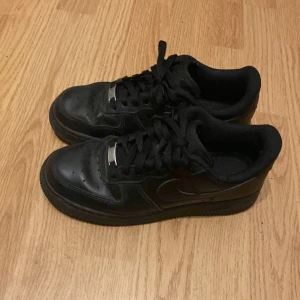 Svarta Nike Air Force 1 sneakers - Klassiska helsvarta Nike Air Force 1 sneakers med låg profil. Skorna har snörning, rund tå och är tillverkade i skinn med en platt sula. Perfekta för en stilren och tidlös look.