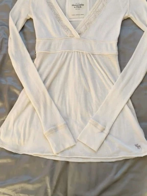 Vit långärmad topp Abercrombie - Säljer en vit långärmad topp från Abercrombie & Fitch med v-ringning och spetsdetaljer vid halsen. Toppen har en lätt utsvängd passform och ribbad struktur, perfekt för en avslappnad men ändå snygg look.